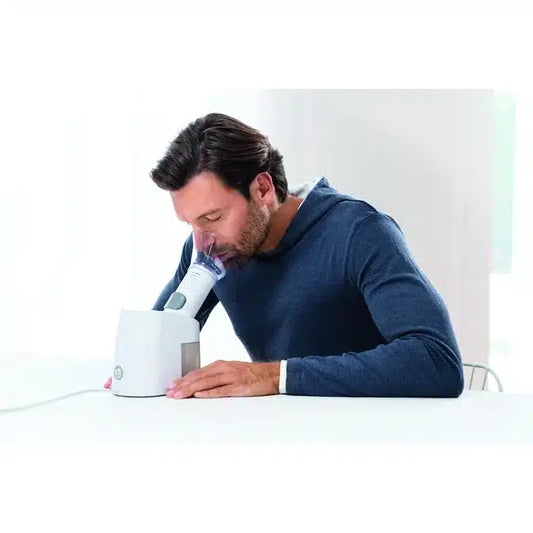 Ein Mann mit braunen Haaren und Bart, in einem blauen langärmeligen Hemd, benutzt den Beurer Dampfvernebler SI 40 Inhalator mit einer Gesichtsmaske an einem weißen Tisch in einem hellen, minimalistischen Raum. Produkt der Beurer GmbH.