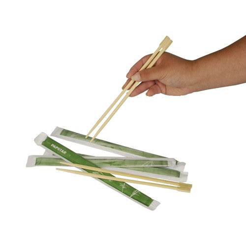 A hand holds wooden PAPSTAR chopsticks "pure" 23 cm over several pairs, each individually wrapped in green paper sleeves. Product: PAPSTAR chopsticks "pure", 50 pairs | Film by PAPSTAR GmbH, depicted on a white background.
