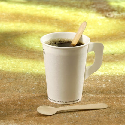 A white paper cup with a handle contains dark coffee and a PAPSTAR wooden coffee spoon "pure", 11 cm from PAPSTAR GmbH as a stirrer; next to it lies another wooden spoon against a blurred yellowish background.