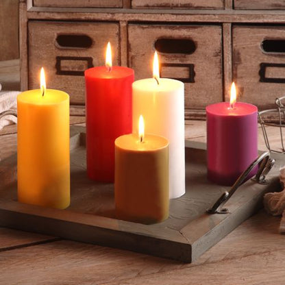 Five PAPSTAR pillar candles Ø 70 mm, 150 mm made of 100% stearin in yellow, red, white, brown, and pink from PAPSTAR GmbH are on a square wooden tray with metal handles on a table, with a wooden drawer unit in the background.