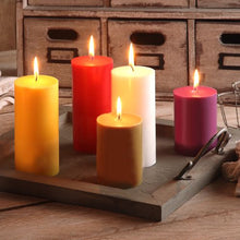 Five burning PAPSTAR pillar candles Ø 70 mm, 100 mm dyed candles from PAPSTAR GmbH made of 100% stearin in yellow, red, white, brown, and pink are placed on a wooden tray with handles on a rustic table in front of small wooden drawers.