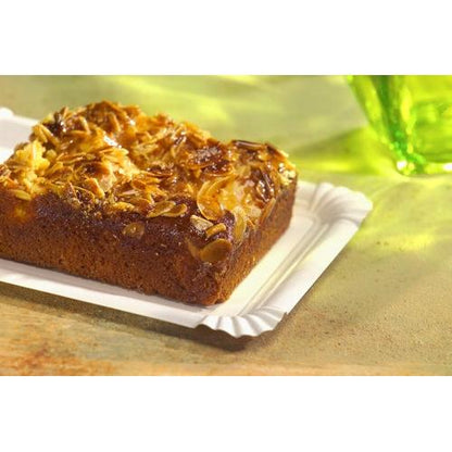 A rectangular piece of almond cake rests on a PAPSTAR GmbH 250 plate, cardboard "pure" square white. In the background, a green glass is blurred.
