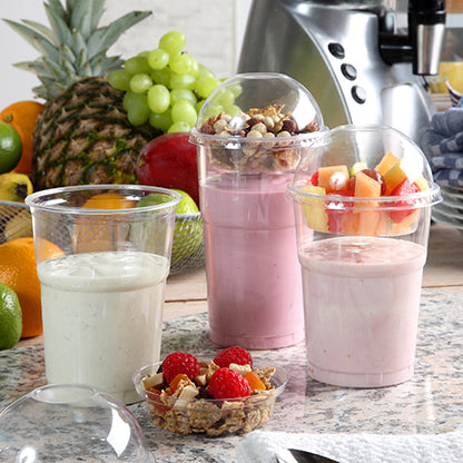 Three smoothies in PAPSTAR GmbH cold drink cup PLA "pure" crystal clear 0.5 l stand on a kitchen counter. Two have fruits and muesli under domed lids. Fresh fruits and a blender can be seen in the background. Package contains 60 pieces.