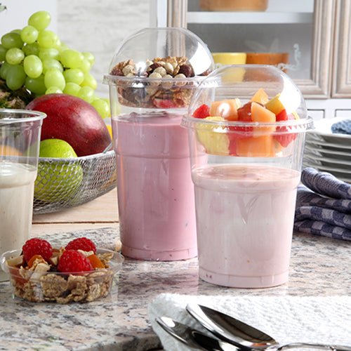 Two yogurt parfaits in plastic cups, each sealed with a Starpak 50 dome lid "Hurricane" PET round crystal clear Ø 9.5 cm, are on a kitchen counter – one with muesli and nuts, the other with fruit. In the background, fresh berries and stacked plates can be seen.