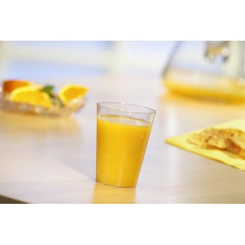 A Starpak 50 drinking cup (PS crystal clear) filled with orange juice stands on a light table, with a blurred plate of orange slices and a yellow napkin with bread or crackers in the background.
