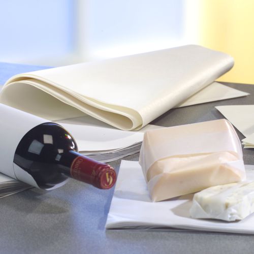 A wine bottle stands on a gray surface, next to PAPSTAR parchment substitute "pure" white wrapping paper (12.5 kg, grease-resistant from PAPSTAR GmbH) and two blocks of cheese, one of which is partially wrapped. The background is slightly blurred.