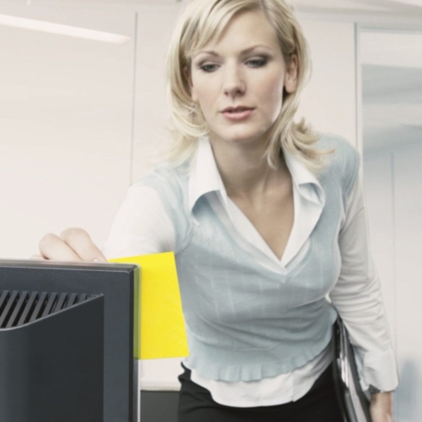 A businesswoman in a modern office sticks a Post-it® Notes Cube 636-B, Yellow, 76 mm x 76 mm, from 3M Germany GmbH on her monitor while holding documents. The self-adhesive notes serve as a reminder aid.