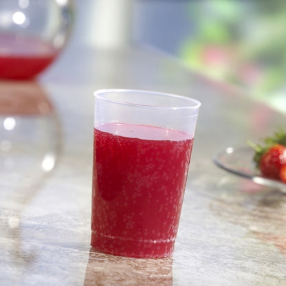 A Starpak 40 reusable drinking cup, translucent unbreakable PP, filled with red fruit punch, stands on a marble surface, with a matching jug of punch and a plate of strawberries blurred in the background.