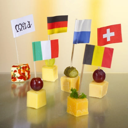 Six cheese cubes are topped with grapes or herbs and decorated for a stylish and eco-friendly presentation with the 8 cm large white deco-picker flags from PAPSTAR GmbH, with each flag labeled "Mild", Germany, Italy, Finland, Switzerland, or Belgium.