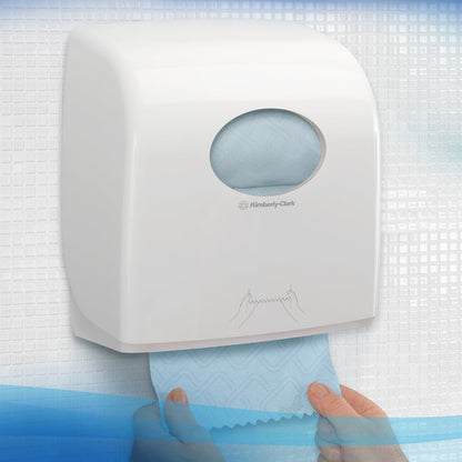 A person pulls a blue Scott® Essential™ towel roll from a white dispenser by Kimberly-Clark GmbH, mounted on a tiled wall with a round window showing the towels inside and an illustration for towel dispensing.