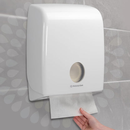 A hand pulls a Kleenex® folded hand towel - interfold from a white wall dispenser by Kimberly-Clark GmbH in a tiled bathroom. The dispenser has a round window that reveals the paper towels inside (15 clips x 186 sheets).