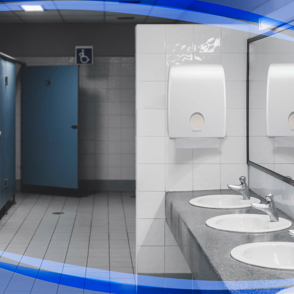 A public restroom with three sinks, a large mirror, blue stall doors - one of them wheelchair accessible - and white tiled floors and walls features a Kimberly-Clark GmbH Aquarius™ dispenser for folded towels above the sinks.
