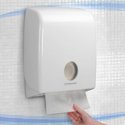A hand pulls a paper towel from a wall-mounted, white Aquarius™ dispenser for folded towels with C-fold from Kimberly-Clark GmbH, installed on a tiled wall.