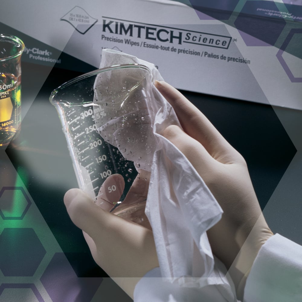 A person with gloves wipes a beaker with Kimtech® Science precision wipes (Kimberly-Clark GmbH, 200 sheets/carton, 15 cartons/carton), which are ideal for sensitive surfaces. A box of laboratory wipes and another beaker are in the background.
