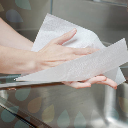 A person dries their hands with Kleenex® Hand Towels - MultiFold / White by Kimberly-Clark GmbH over a metal sink, with semi-transparent water droplets in the background creating a fresh, modern washroom feel.