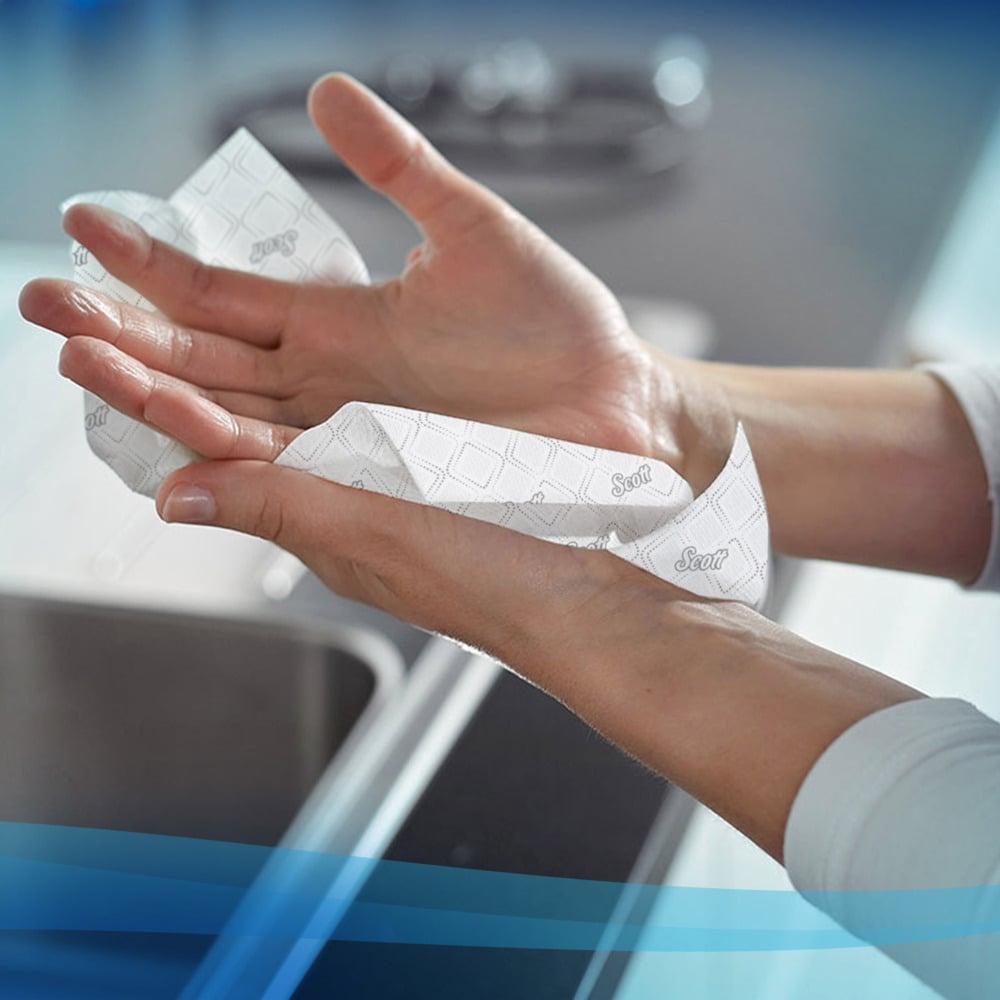Someone dries their hands at the sink with a white, patterned Scott® Hand Towels - Narrow-Fold paper towel by Kimberly-Clark GmbH, which highlights hygienic hand drying alongside blurred appliances and a gentle blue gradient.