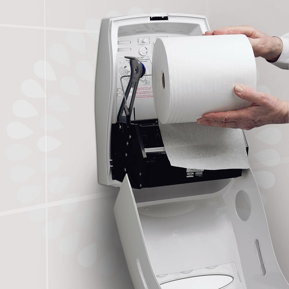 A person loads a Kleenex® Ultra™ hand towel roll from Kimberly-Clark GmbH into a white, open wall dispenser in a toilet to ensure hygienic hand drying. Instructions are printed on the lid of the dispenser.