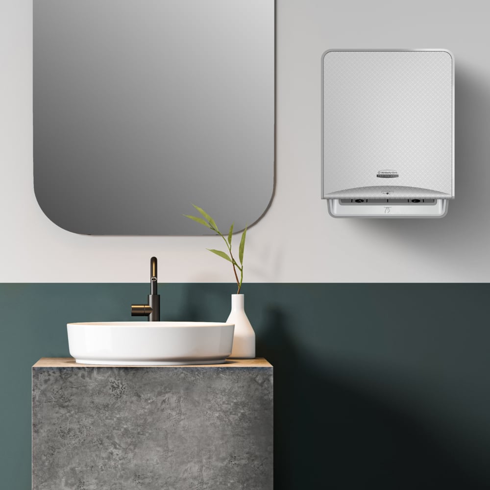Modern bathroom with rectangular mirror, white sink on a vanity with concrete texture, black faucet, white vase with green branches, and a wall-mounted dispenser for Kleenex® Roll Towels from Kimberly-Clark GmbH.