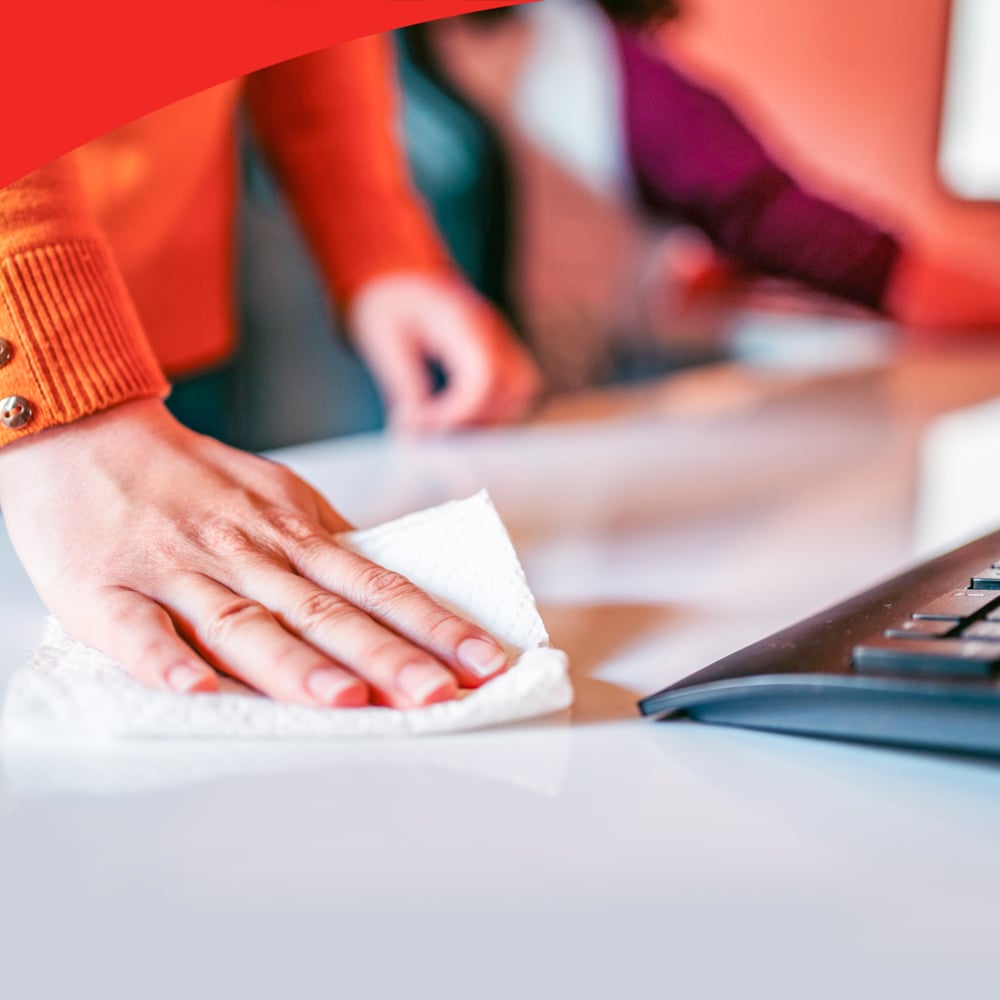 A person in an orange sweater cleans a white desk near a black keyboard with WypAll® X60 General Clean white quarter-folded cloths (12 bags x 76 sheets) from Kimberly-Clark GmbH, with another person visible in the background out of focus.