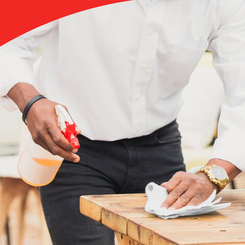 A person with a white shirt and a gold watch cleans a wooden table with a cloth and a spray bottle, possibly using Kimberly-Clark WypAll® L20 General Clean large roll (2 rolls x 1085 sheets). In the upper part of the image, a curved red section is visible.