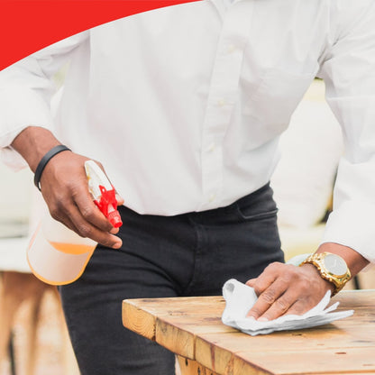 A person with a white shirt and a gold watch cleans a wooden table with a cloth and a spray bottle, possibly using Kimberly-Clark WypAll® L20 General Clean large roll (2 rolls x 1085 sheets). In the upper part of the image, a curved red section is visible.