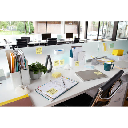 A modern office table with an open planner, closed laptop, pen, coffee cup, plant, folders, and 3M Germany GmbH Post-it® Notes Yellow (51x76mm, 100 sheets/pad), with large windows with views and more desks behind.