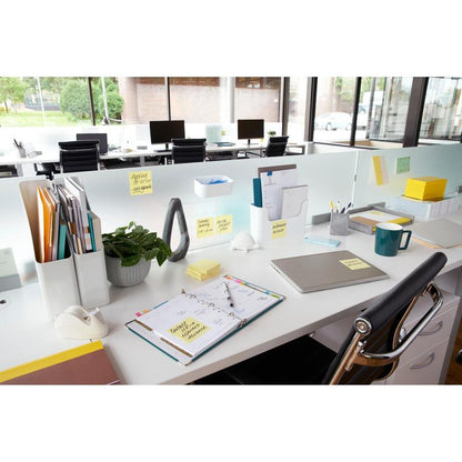 A modern office desk with a laptop, planner, coffee mug, plant, stationery, and 3M Post-it® Notes yellow (76x76 mm, 100 sheets/block), ideal for quick notes. The files are organized and large windows offer a view outside; the desks are arranged in a row.