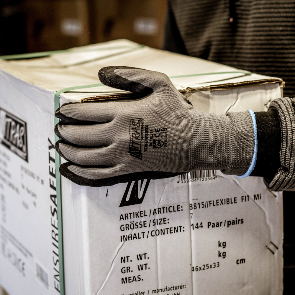A person wearing Nitras Nylotex nylon gloves with latex coating from AS Arbeitsschutz GmbH holds a large white box with German and English labeling in their hand; only their upper body and arm are partially visible.