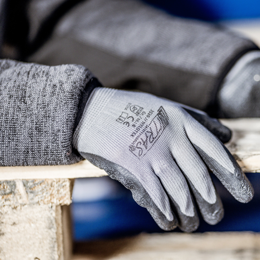 A close-up shows a person wearing a textured, gray Nitras Nylotex nylon glove with latex coating from AS Arbeitsschutz GmbH and resting their hand on a wooden board. Markings and text are visible on the back; the background is slightly blurred.