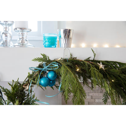 Ein mit Tannengirlanden, blauen Ornamenten, Bändern und Lichtern geschmückter Kaminsims wird von silbernen und blauen Kerzenhaltern überragt, die mit 3M Command™ Haken M transparent (17091CLR) rückstandsfrei aufgehängt wurden. Im Hintergrund ist ein Fenster zu sehen.