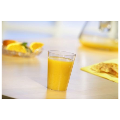 A Starpak 50 drinking cup, PS crystal clear by Starpak, filled with orange juice, stands on a light table. Behind it are orange slices on a plate and a blurred yellow object, possibly food, on a yellow napkin.