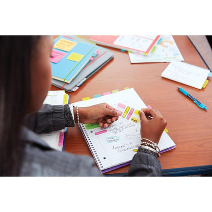 A person uses the 3M Deutschland GmbH Post-it® Z-Notes Dispenser (PRO, black) with Super Sticky Z-Notes (yellow, 76x76 mm, 90 sheets/block) in a notebook on a wooden table, surrounded by folders, papers, and a pen.
