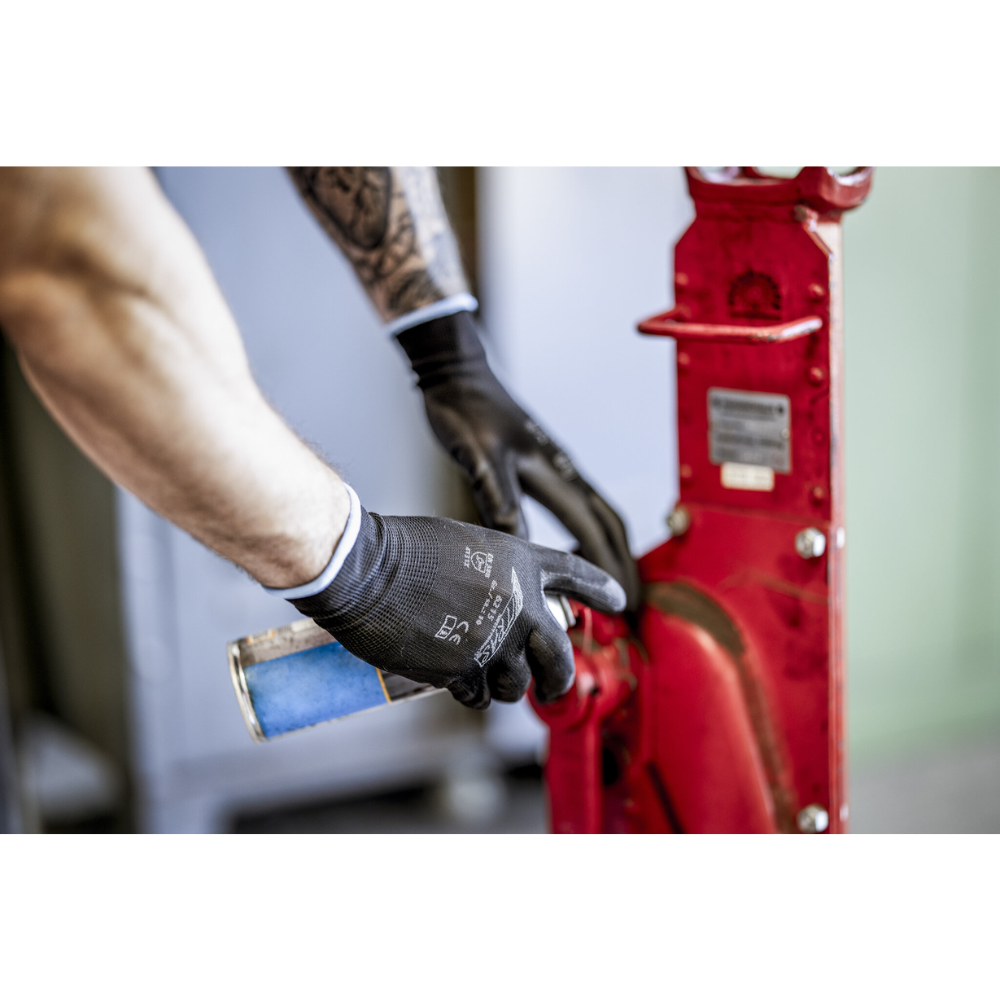 A person with NITRAS nylon gloves, black, PU-coated from AS Arbeitsschutz GmbH cleans or lubricates a red metal machine part with a blue spray can. Only arms and hands are visible.