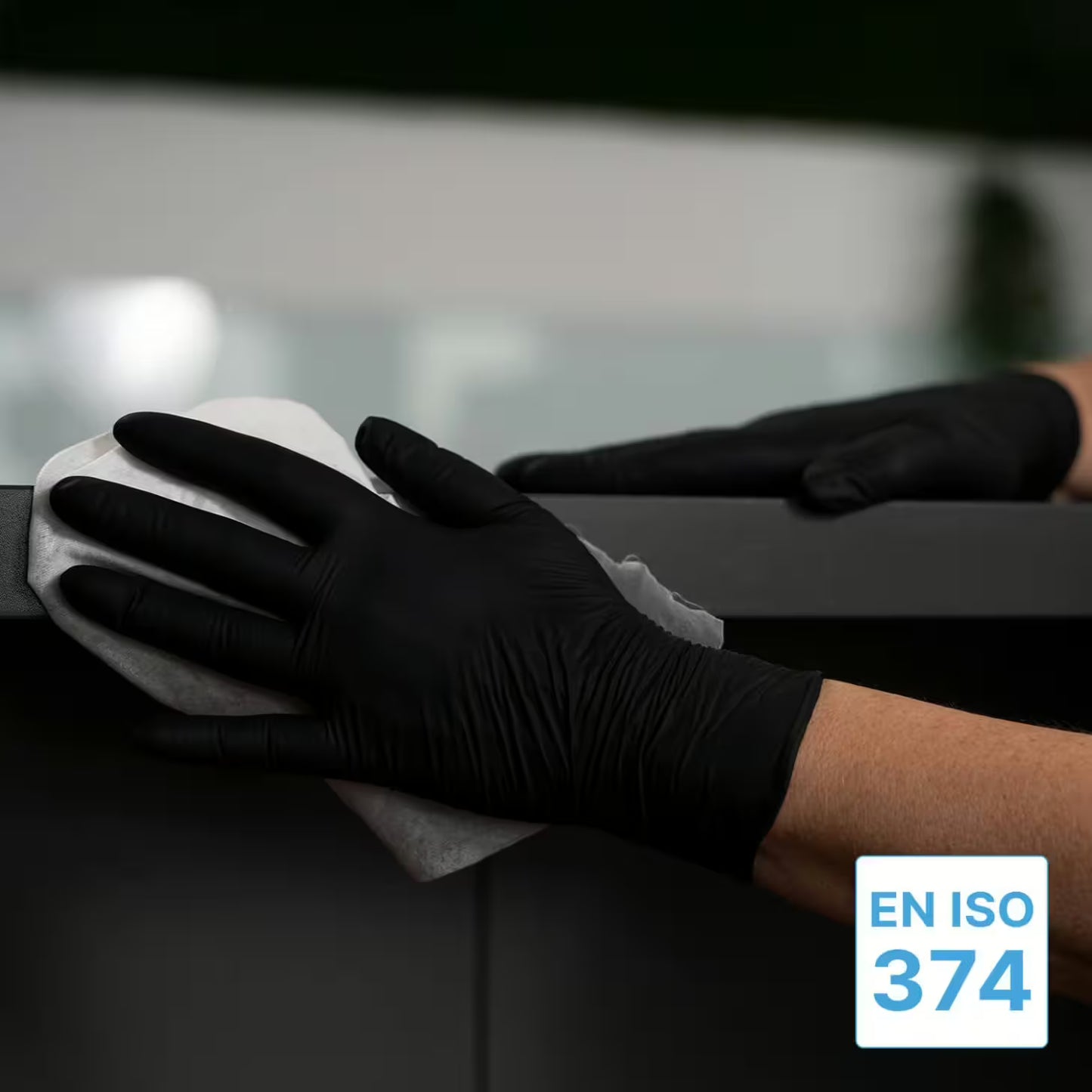 A person in ARNOMED NITRIL XTRA PURE BLACK, skin-friendly & accelerator-free nitrile disposable gloves from ARNOWA GmbH, wipes a dark surface with a white cloth. In the lower right corner, an "EN ISO 374" certification label is depicted.
