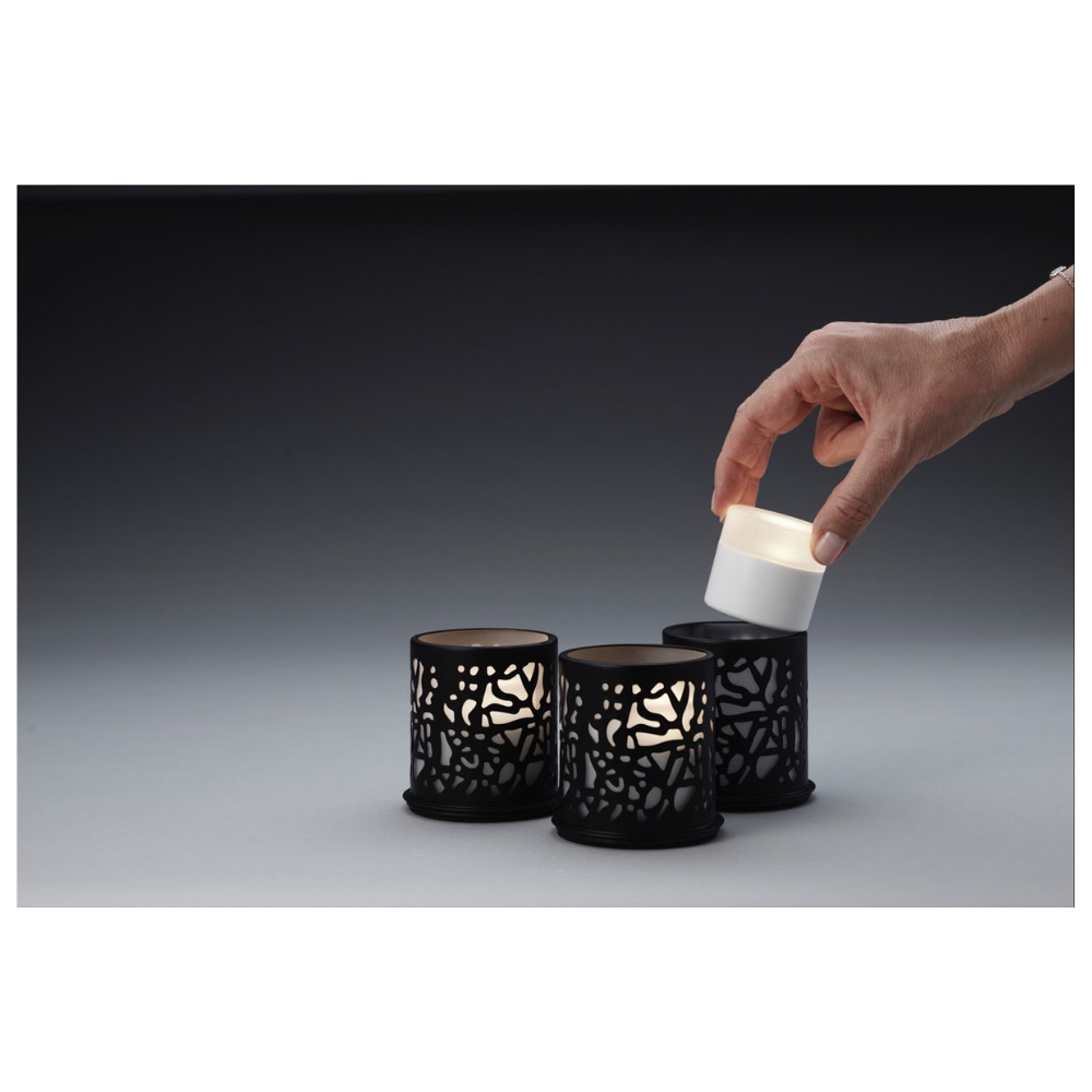 A hand places a small cylindrical candle into one of three Twine 75 x Ø 75 mm Black candle holders from Duni GmbH, which are adorned with elaborate cut-outs and are ideal for modern design. The table decoration is placed against a gradient background from dark to light gray.