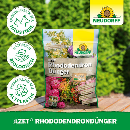 A bag of Azet Rhododendron Fertilizer from the Neudorff Shop lies on a wooden surface outdoors in front of green hedges. The packaging made of bioplastic states that it is organic, pet-friendly, and ideal for rhododendrons and azaleas.