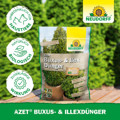 A bag of Neudorff Shop Azet Buxus & Ilex Fertilizer, an organic fertilizer for boxwood and holly, stands outdoors on a table. The packaging indicates its safety for pets and natural ingredients, while lush green shrubs are visible in the background.