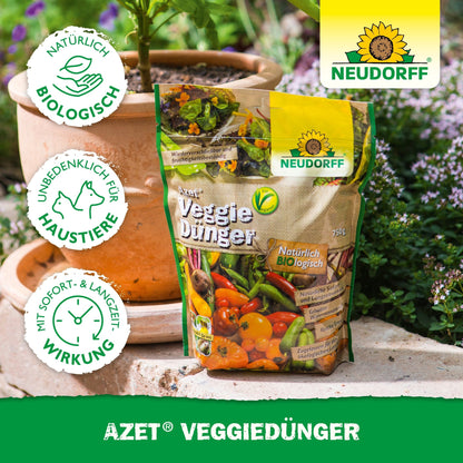 A bag of Neudorff Shop Azet VeggieFertilizer stands on a wooden surface outdoors next to a terracotta pot with green plants. The packaging features vegetable images and certification logos.