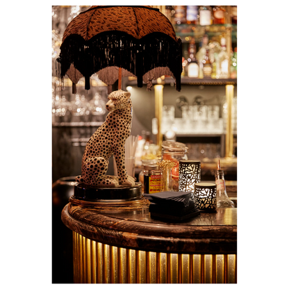 A dimly lit bar with a leopard statue on the counter under a fringed lampshade impresses with modern design. Bottles and napkins surround a chic candle holder Twine 75 x Ø 75 mm Black from Duni GmbH, creating an inviting table decoration next to a metal lantern.
