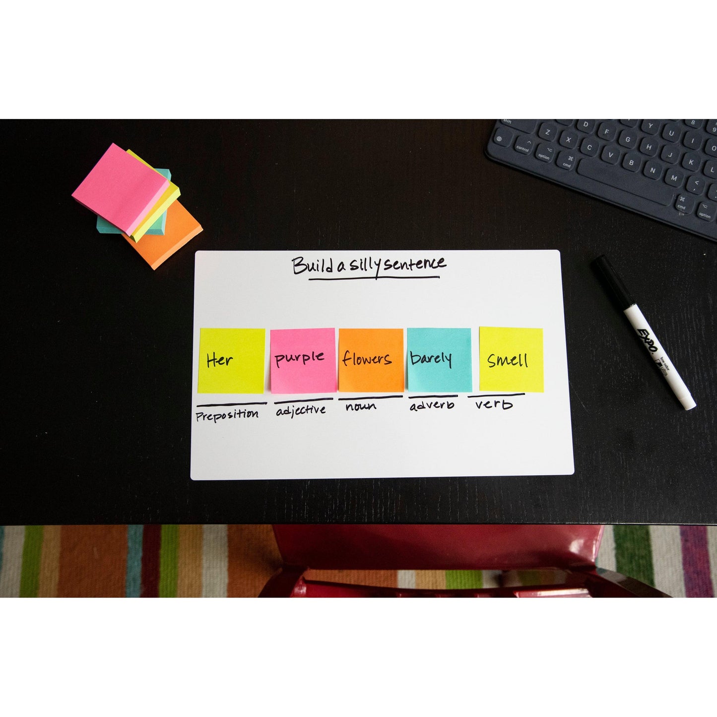 A Post-it® note pad (76mm x 127mm, 100 sheets, 100% PEFC) from 3M Deutschland GmbH lies on a black desk with notes labeled with preposition, adjective, noun, adverb, and verb; also pictured are a marker and a keyboard.