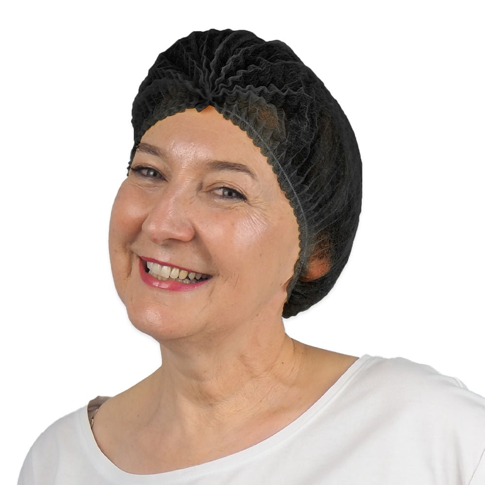 A smiling older woman wears a black ARNOMED COVER PP CLIP CAP disposable clip cap made of PP fleece (52cm, ARNOWA GmbH) and a white shirt, slightly turned to the side against a simple white background.