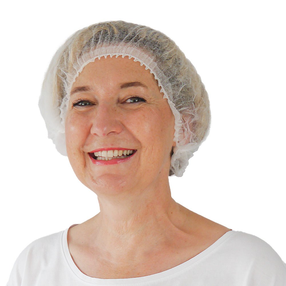 A middle-aged woman smiles at the camera. She is wearing a white ARNOMED COVER PP CLIP CAP disposable clip cap made of PP fleece (52cm) from ARNOWA GmbH, paired with a simple white top against a white background, emphasizing her commitment to hygiene protection.