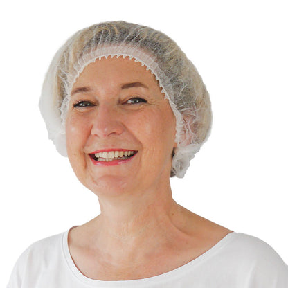 A middle-aged woman smiles at the camera. She is wearing a white ARNOMED COVER PP CLIP CAP disposable clip cap made of PP fleece (52cm) from ARNOWA GmbH, paired with a simple white top against a white background, emphasizing her commitment to hygiene protection.