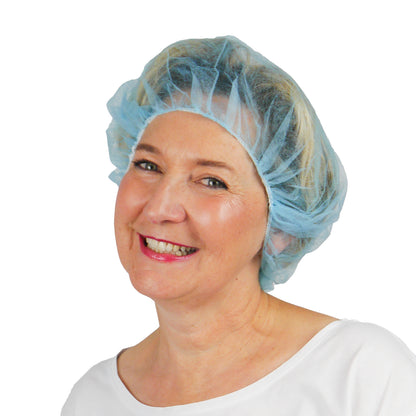 A woman in a light blue ARNOMED COVER PP-BARETTHAUBE bouffant cap made of PP non-woven fabric by ARNOWA GmbH and a white top smiles in front of a plain white background, highlighting this hygienic accessory for the hygiene sector.