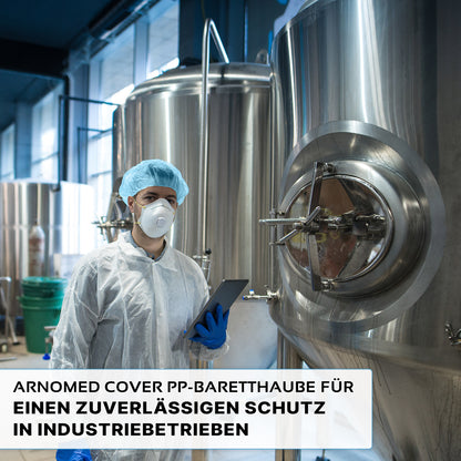A person wearing an ARNOMED COVER PP-BARETTHAUBE bouffant cap made of PP non-woven fabric by ARNOWA GmbH and a mask stands in front of stainless steel tanks in an industrial hygiene area holding a tablet. The German text highlights reliable protection for the industry.