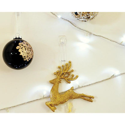 A golden glitter reindeer ornament hangs on a white wall, securely attached with a transparent Command™ Mini Hook, 17006CLR from 3M Deutschland GmbH, next to a black ornament with gold details and white string lights.