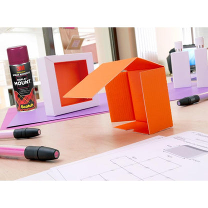 A table with an orange cardboard model, markers, 3M™ Scotch® DisplayMount™ adhesive spray (400 ml, 1 can/package) from 3M Deutschland GmbH, white boxes, and technical drawings in a bright indoor workspace for professional use.