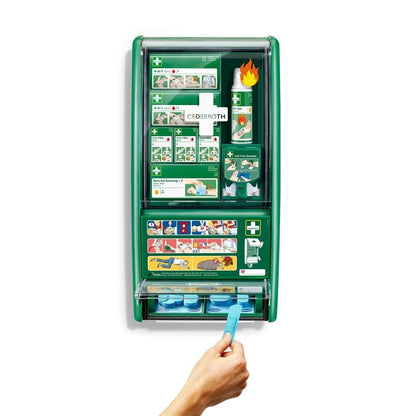 A green Cederroth first aid & burn station from Orkla Wound Care AB is mounted on a white wall, stocked with bandages, burn gel, eye wash, and more. A hand pulls a blue adhesive bandage from the bottom dispenser of the 1-pack box.