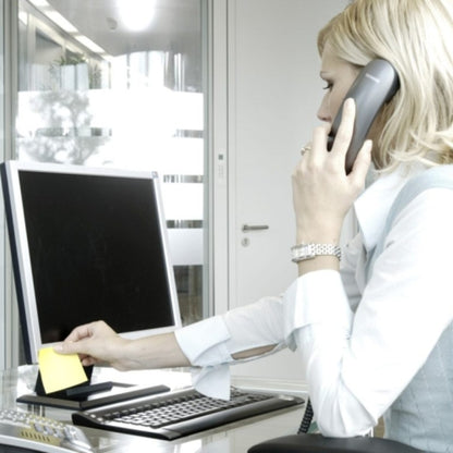 A blonde person at a desk places a 76 mm x 76 mm yellow Post-it® Notes Cube 636-B from 3M Germany GmbH (450 sheets, PEFC) on the monitor while on the phone. Keyboard, mouse, and computer are also on the table.