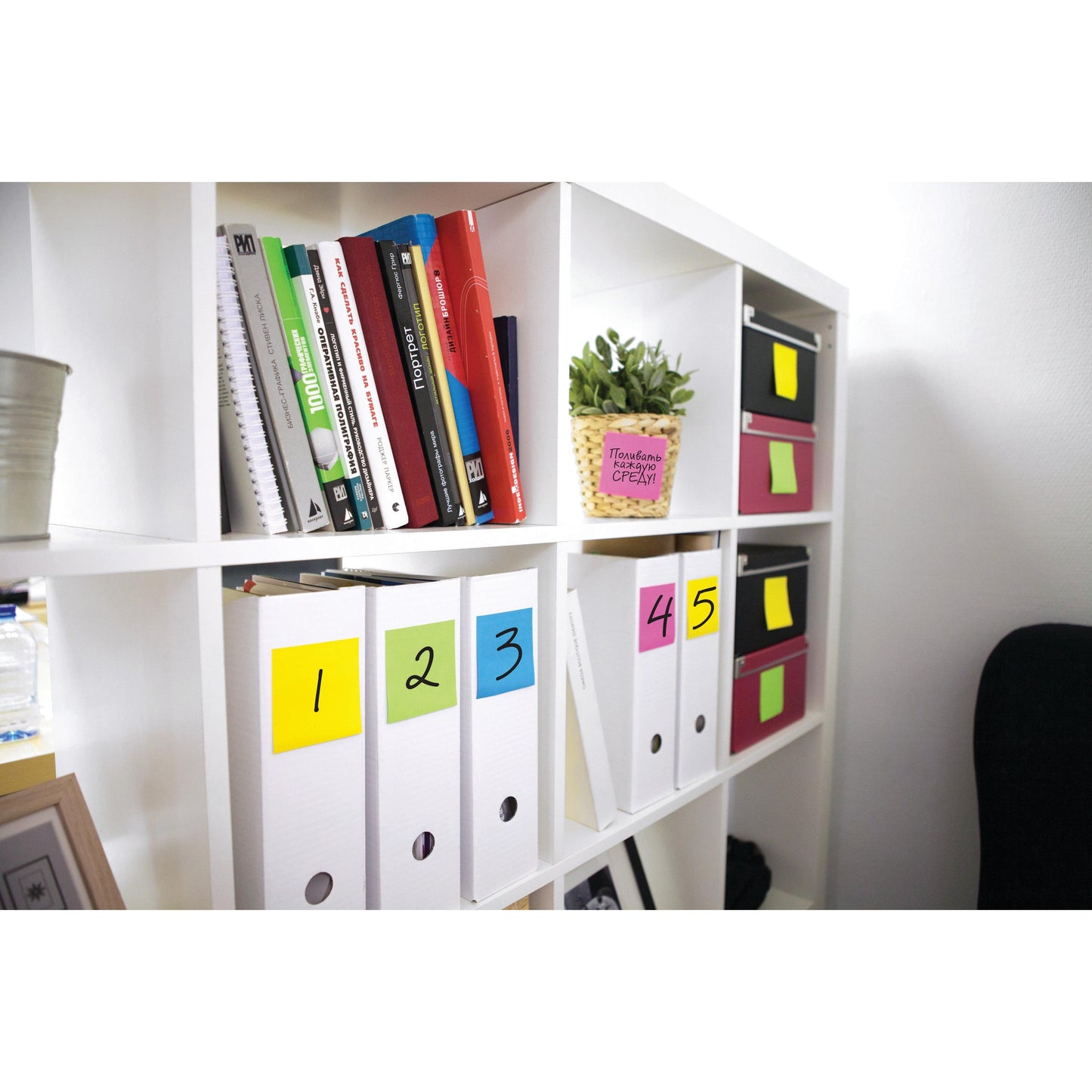 On a white shelf are books, a potted plant, white folders with self-adhesive notes in the format 76 mm x 76 mm, pink and black boxes, and a Post-it® Notes Cube 636-B in Yellow from 3M Germany GmbH (76x76mm, 450 sheets/block).